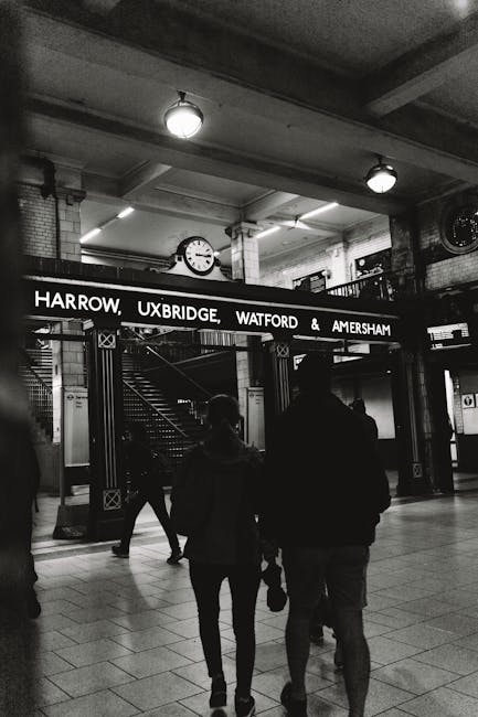 Black and white photograph of the entrance to Harrow, Uxbridge, Watford, and Amersham railway stations at a busy underground station. The scene shows two people walking side by side towards the station entrance, which has a large illuminated sign displaying the station names. The individuals appear to be carrying small bags, and are dressed in casual clothing appropriate for urban travel. The background reveals the station's interior with tiled floors, brick walls, and the station's clock mounted above the entrance. Overhead lighting illuminates the area, highlighting the structural beams and the staircase visible behind the sign. This setting is related to home relocation or moving logistics, emphasizing the importance of transport and transfer of belongings during moving services offered by companies like Uxbridge Man and Van, as suggested by the website information.
