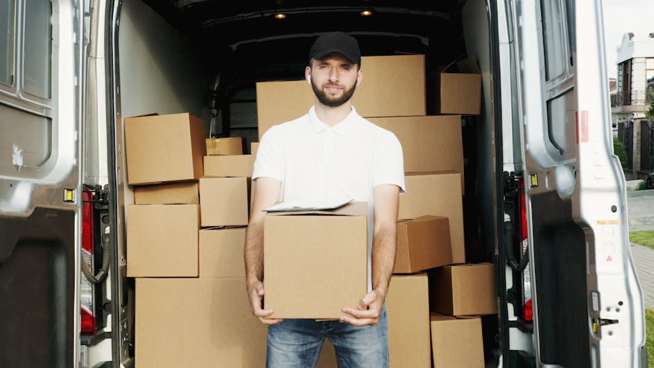 A man wearing a black cap, white shirt, and jeans stands inside the open rear of a white moving van, holding a large cardboard box with both hands. The van's interior is filled with neatly stacked cardboard boxes of various sizes, some wrapped in plastic, others unwrapped, ready for a home relocation. The boxes are arranged on the van's floor and against the walls, indicating an organized packing process typical of furniture transport and packing during house removals. Outside the van, a paved driveway and parts of residential buildings are visible, suggesting the location is a suburban neighbourhood. The lighting is natural daylight, illuminating the scene clearly. Uxbridge Man and Van, a professional removals service, is preparing for a house removal or moving process, demonstrating the loading phase of packing and transport logistics.