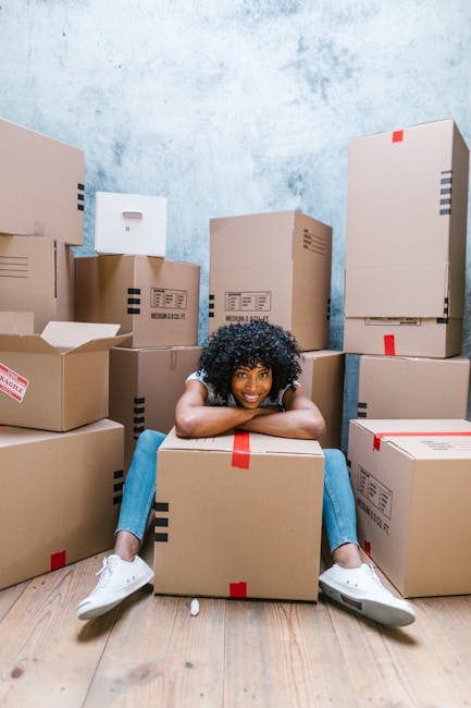 A young woman with curly black hair, smiling and sitting on a large cardboard box inside a room with a textured, light blue wall. She is wearing a white T-shirt, blue jeans, and white sneakers. Surrounding her are numerous packing boxes of various sizes, some sealed with red tape and others open, revealing packing paper and internal contents. The boxes are stacked against the walls and on the wooden floor, indicating a home relocation or move-in process. The scene captures the packing stage of house removals, with boxes prepared for transport. This visual aligns with the context of furniture transport, packing and moving, and loading activities typical of professional removals services, such as those offered by Uxbridge Man and Van.