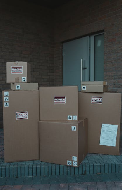 A collection of large cardboard boxes stacked outside a building, positioned on a brick pavement near a metal door. The boxes are sealed with packing tape and feature labels indicating that they are fragile. Some boxes have blue stickers with handling symbols, such as arrows for upright orientation and recycling icons. The boxes vary in size, with one placed on top of another and others arranged side by side, ready for transport. The environment appears to be an exterior area adjacent to a brick wall and a blue metal door, with natural daylight illuminating the scene. These boxes are part of a home relocation process managed by Uxbridge Man and Van, involving packing and loading items for moving or furniture transport as part of their removals services.