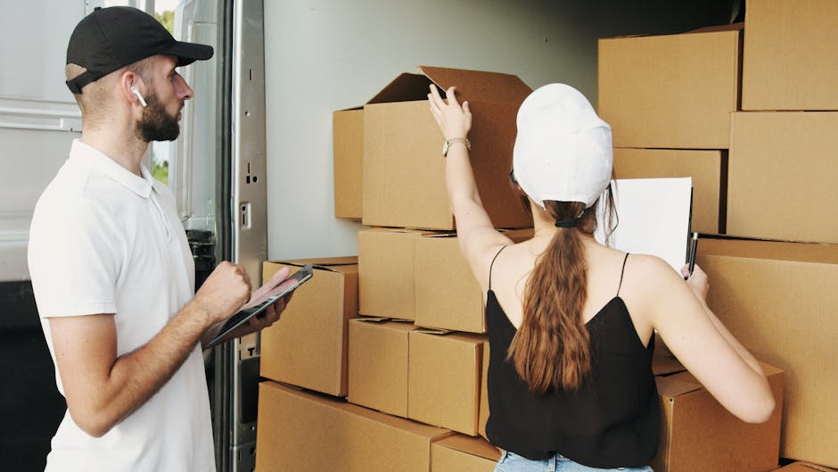 A young woman with curly black hair, smiling and sitting on a large cardboard box inside a room with a textured, light blue wall. She is wearing a white T-shirt, blue jeans, and white sneakers. Surrounding her are numerous packing boxes of various sizes, some sealed with red tape and others open, revealing packing paper and internal contents. The boxes are stacked against the walls and on the wooden floor, indicating a home relocation or move-in process. The scene captures the packing stage of house removals, with boxes prepared for transport. This visual aligns with the context of furniture transport, packing and moving, and loading activities typical of professional removals services, such as those offered by Uxbridge Man and Van.