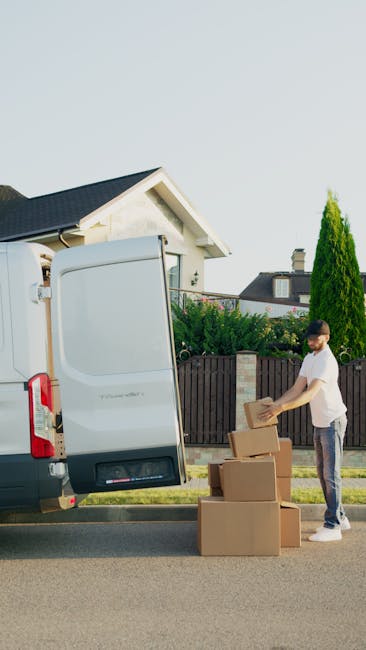 A man dressed in a maroon polo shirt, red shorts, and black work boots is outside, pushing a red-handled hand truck loaded with large cardboard boxes, one marked 'LARGE' and another smaller box on top